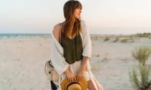Kobieta w letnich stylizacjach boho jesień na plaży, z kapeluszem w dłoni i plecioną torbą.