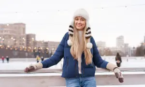 Uśmiechnięta dziewczyna w granatowej kurtce i futrzanej czapce z pomponami, zastanawia się, jaka czapka do granatowej kurtki pasuje najlepiej na lodowisko.