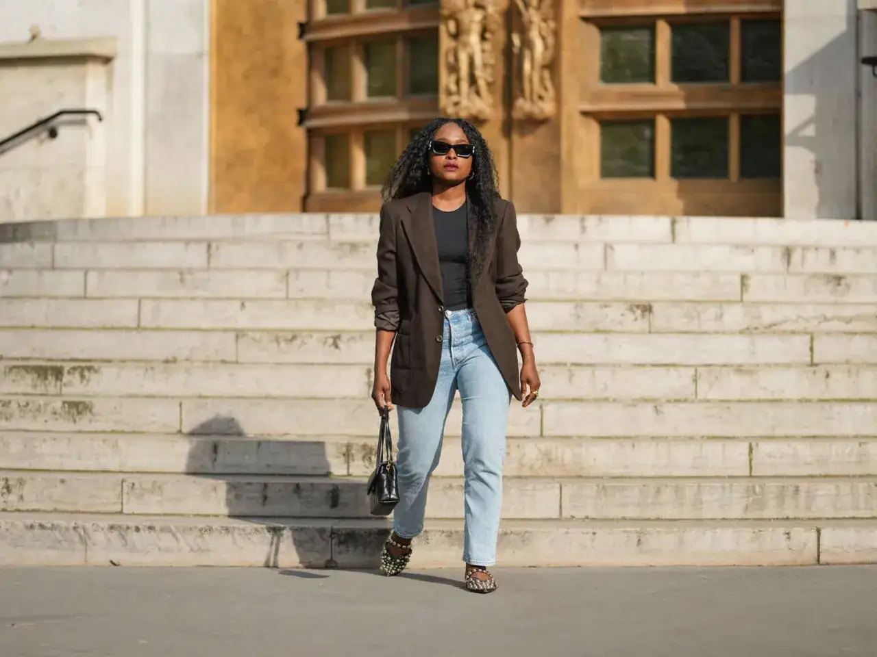 Eleganckie stylizacje z jeansami: kobieta w brązowej marynarce i jasnych jeansach, z czarną torebką i ozdobnymi sandałkami, idzie po schodach.