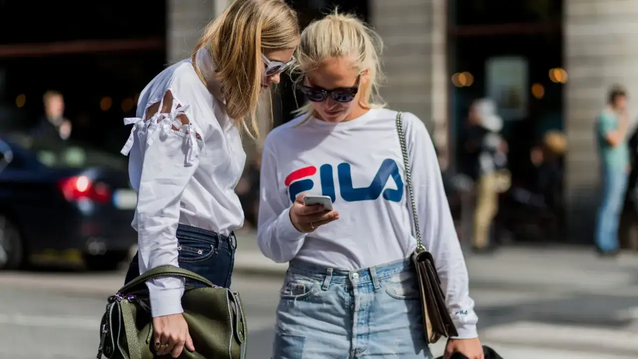 Dwie kobiety w stylu lat 90. moda damska: jedna w białej koszuli z wycięciami na rękawach, druga w bluzie Fila i jeansowych szortach.